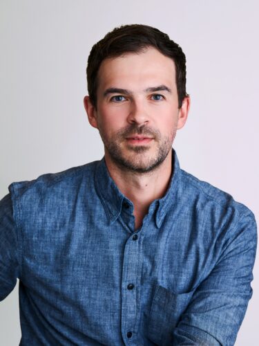 Man with short dark hair and beard wearing a blue button-up shirt, posed in front of a plain background.