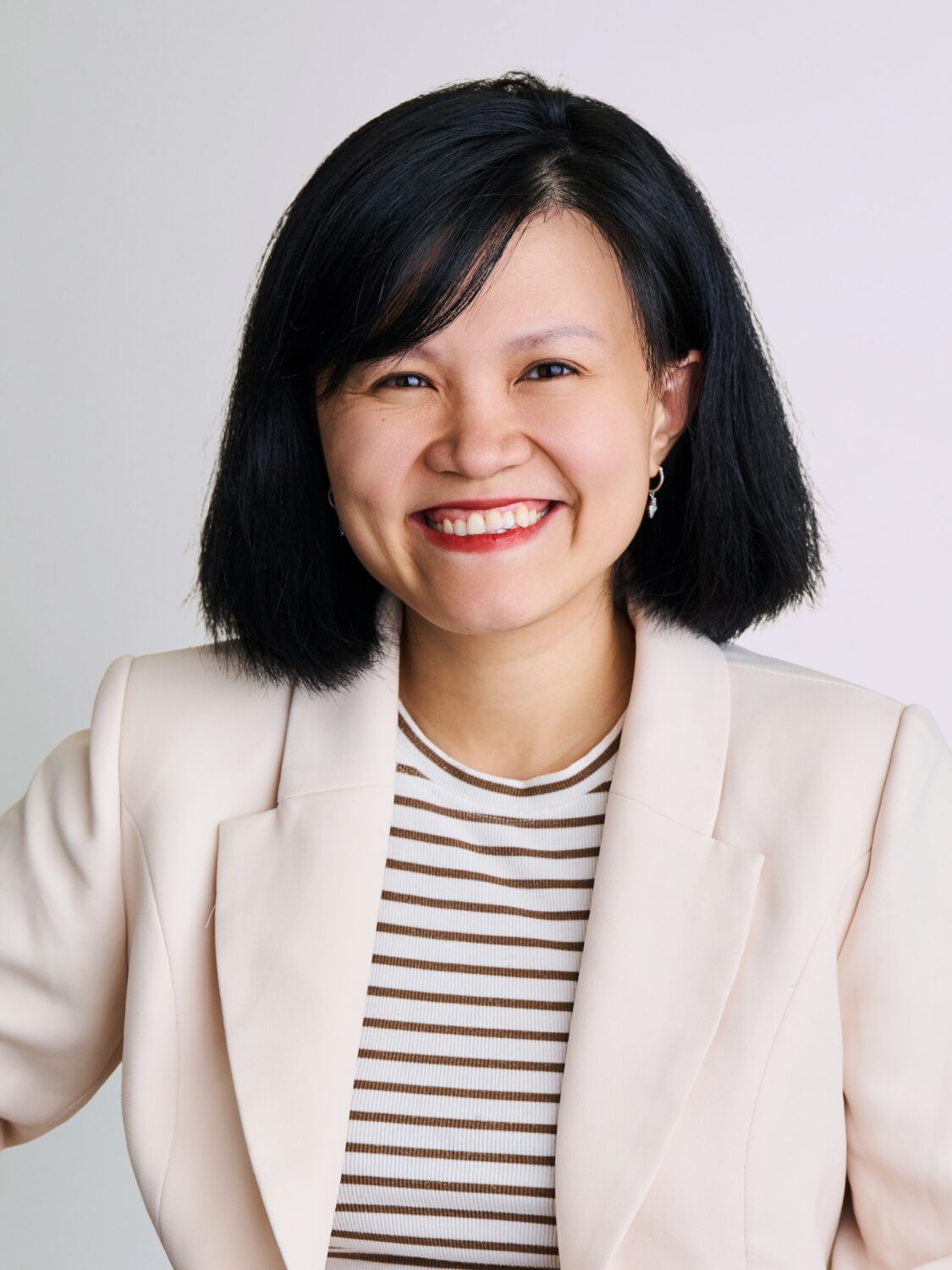 Woman with short black hair, smiling, wearing a light blazer and striped top against a light background.