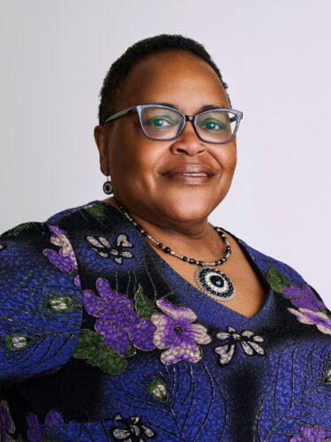 Smiling woman in glasses and a floral-patterned top with matching necklace, posing against a plain background.
