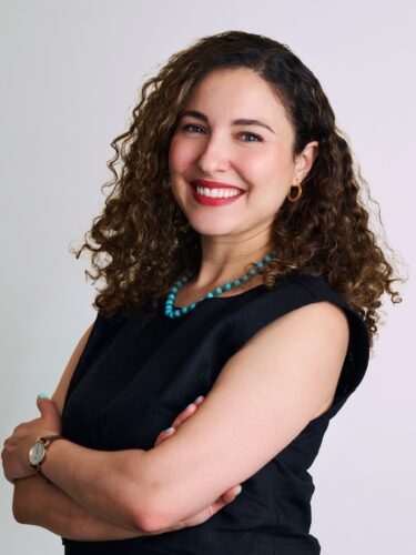 Woman with curly hair in a black top, smiling with arms crossed against a plain background.