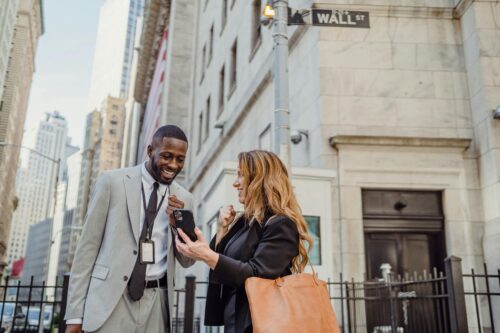 Two professionals smile and look at a phone together on a bustling New York city street near a Wall Street sign.