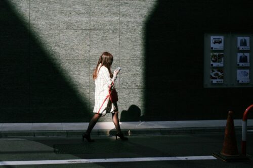 A woman in a white coat walks on a sunlit street, looking at her phone, with a shadowy wall behind her—perhaps reading the latest Meta Antitrust updates from the Omidyar Network.