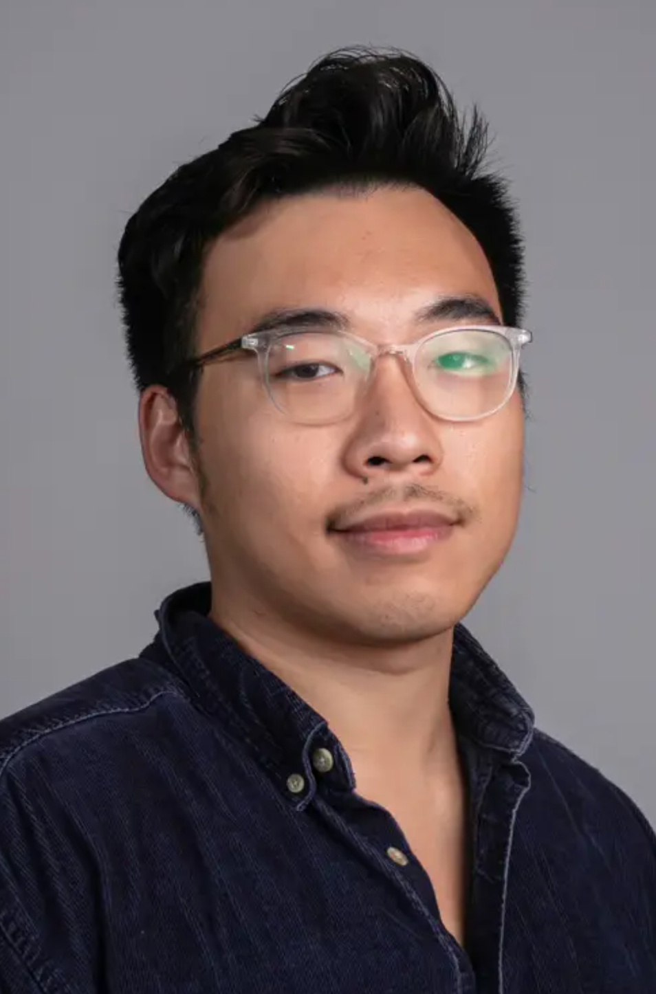 A man with glasses and a dark shirt poses against a plain gray background, looking slightly toward the camera.