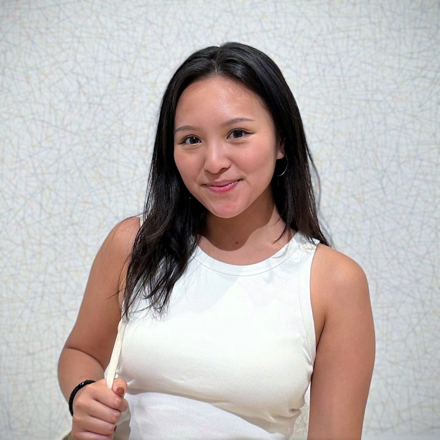 Smiling woman in a white sleeveless top stands against a light, abstract patterned background.