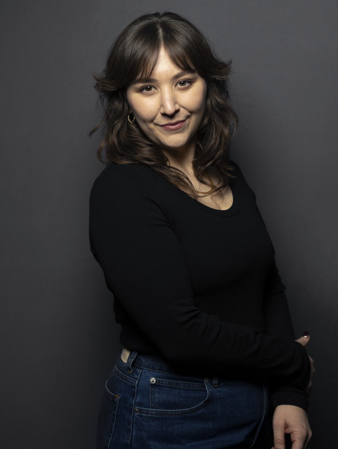 Lee V. Gaines, a woman with wavy brown hair in a black top and jeans, stands against a dark gray background, smiling gently.