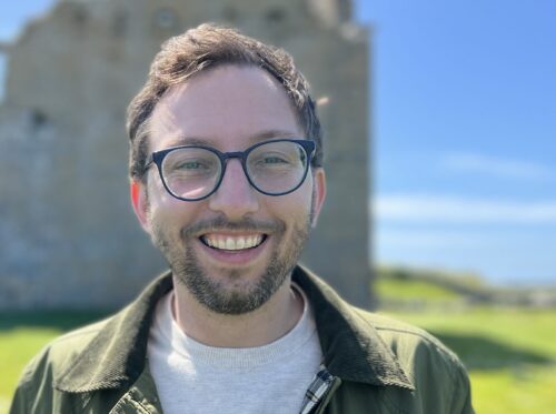 Man with glasses smiling outdoors on a sunny day, with a stone building and blue sky in the background.