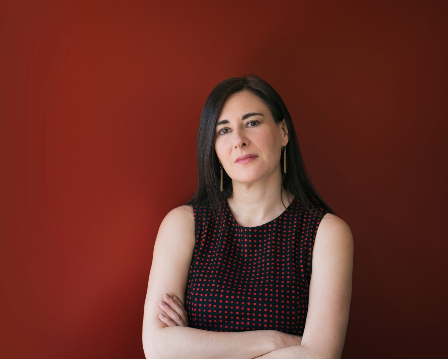 Sharon Weinberger, a defense journalist with long dark hair in a sleeveless top, stands against a plain red background, arms crossed.