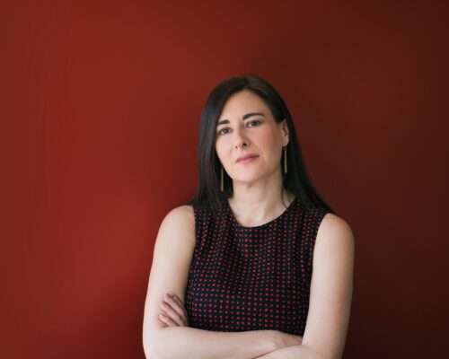 Sharon Weinberger, a defense journalist with long dark hair in a sleeveless top, stands against a plain red background, arms crossed.