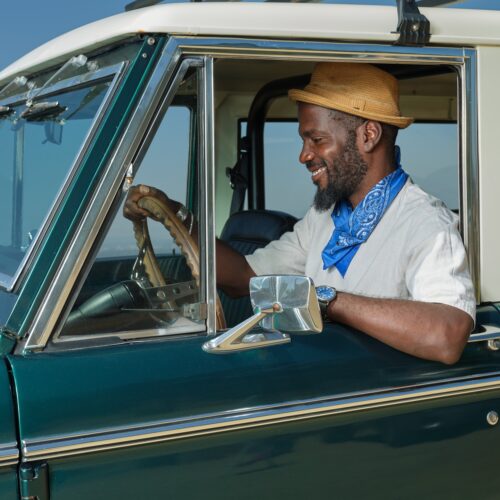 Sonari Batubo Glinton, smiling in a straw hat and bandana, sits in a vintage green car with the window down.