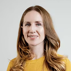 Sylvia Varnham O’Regan, with long light brown hair, smiles warmly in a yellow sweater against a plain light background.