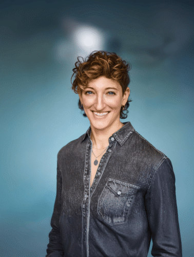 Smiling person with short curly hair in a denim shirt, standing against a blurred blue background.