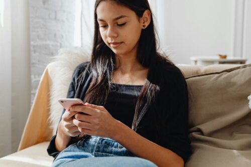 A young woman sits on a couch, looking at her smartphone with a neutral expression.